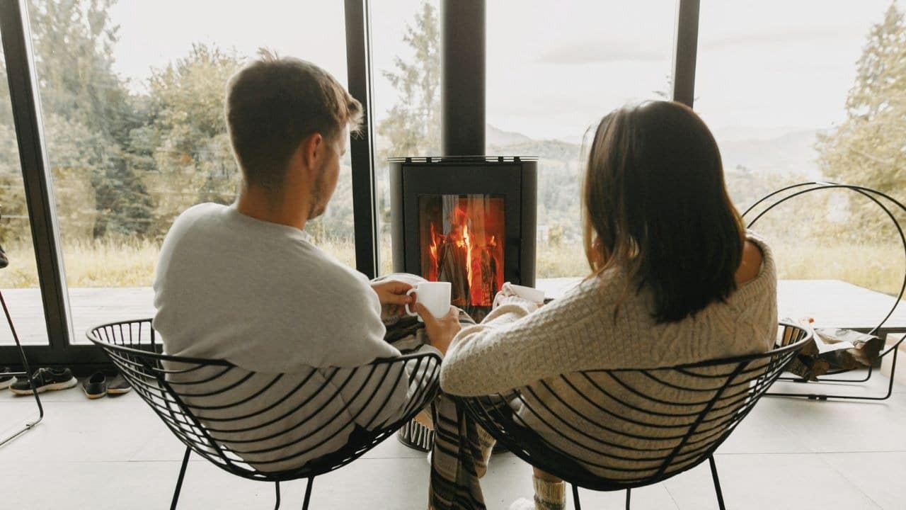 A man and woman sit in wire chairs facing a wood-burning stove with a fire.