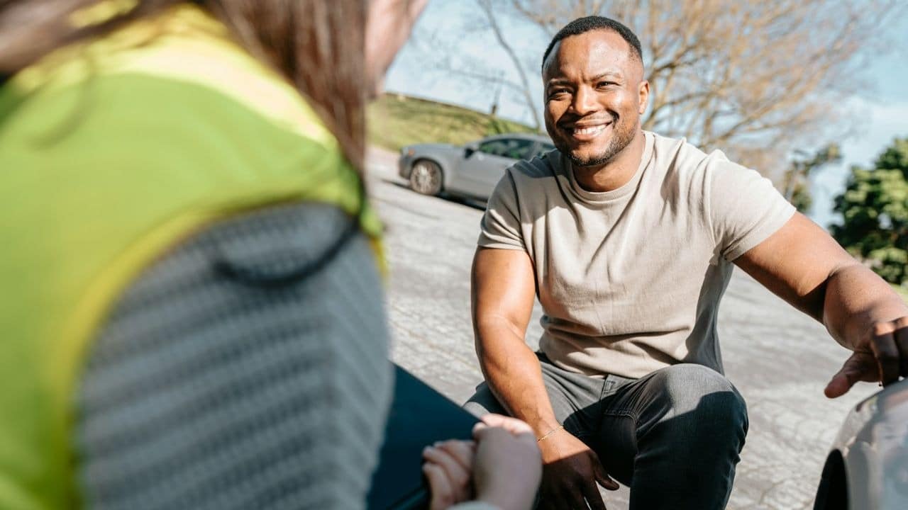A man smiles while kneeling on a paved surface and talking to a person nearby.