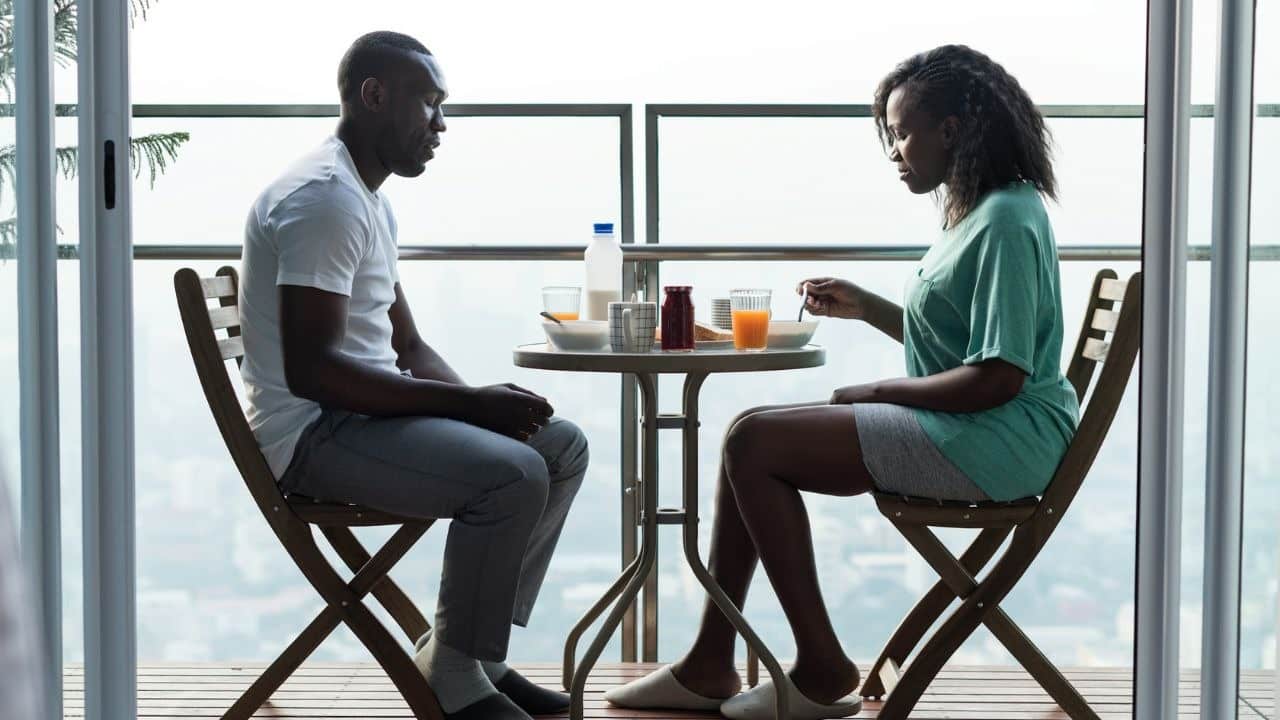 A man and woman sit at a small balcony table with breakfast food between them.
