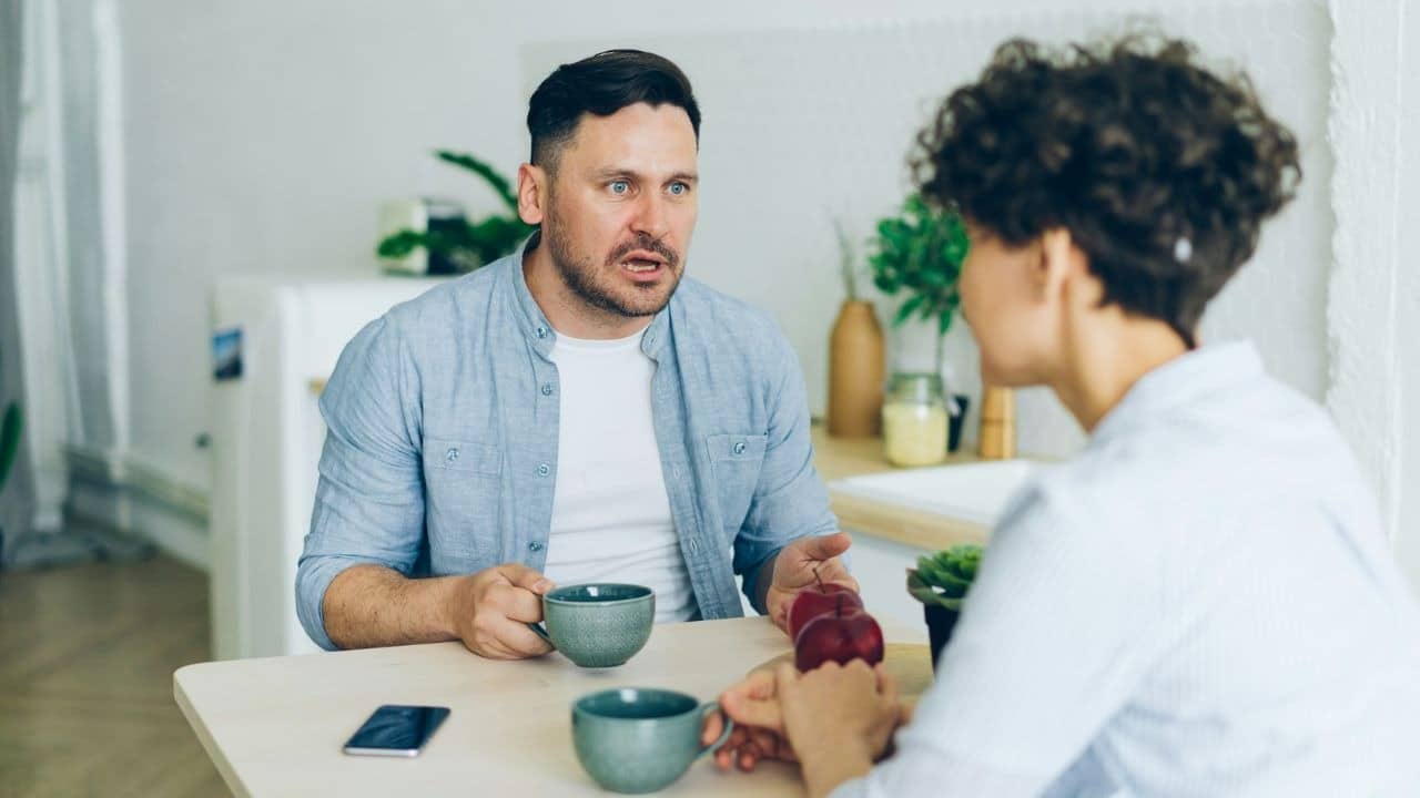 A man speaks while holding a mug at a table across from a woman.