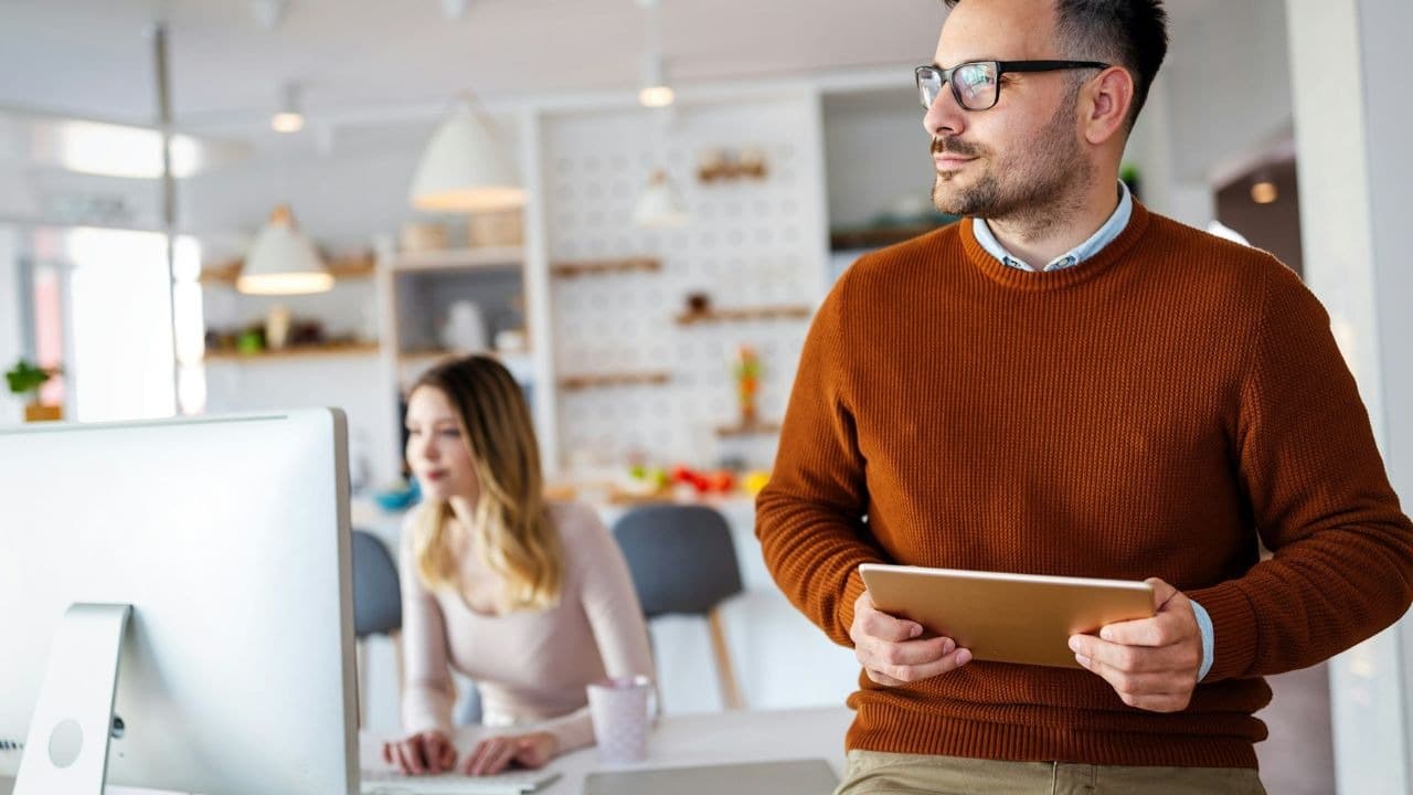 A man holds a tablet while a woman works at a computer in the background.