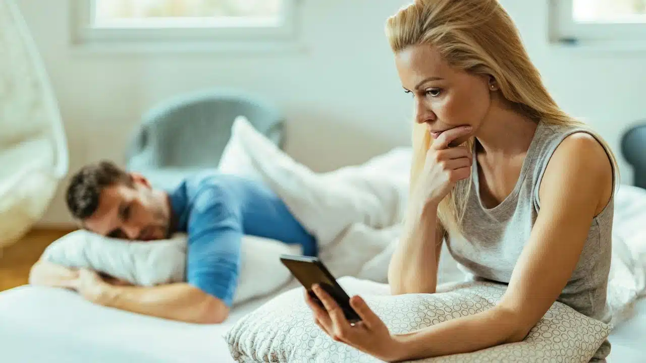 A woman looks at a phone while a man sleeps on a bed behind her.