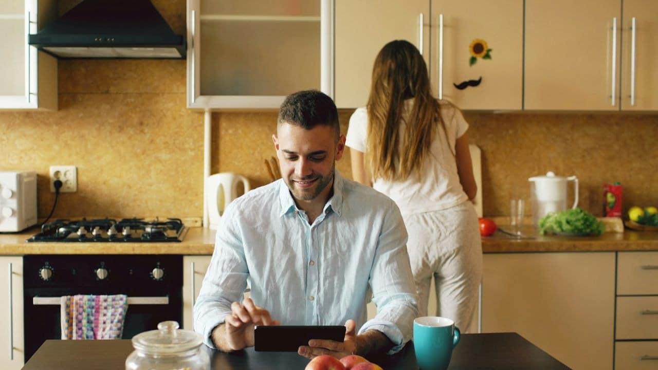 A man uses a tablet at a table while a woman works in the kitchen behind him.