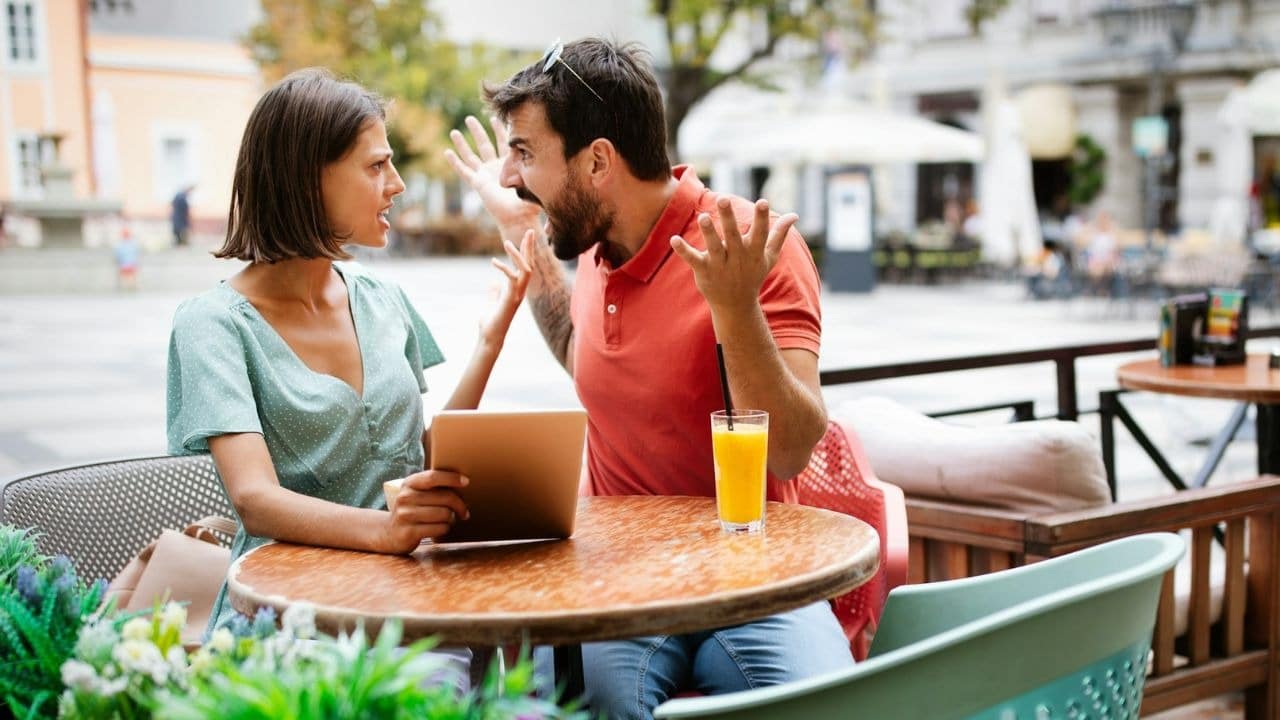 A man shouts with raised hands at a woman sitting across from him at an outdoor table.