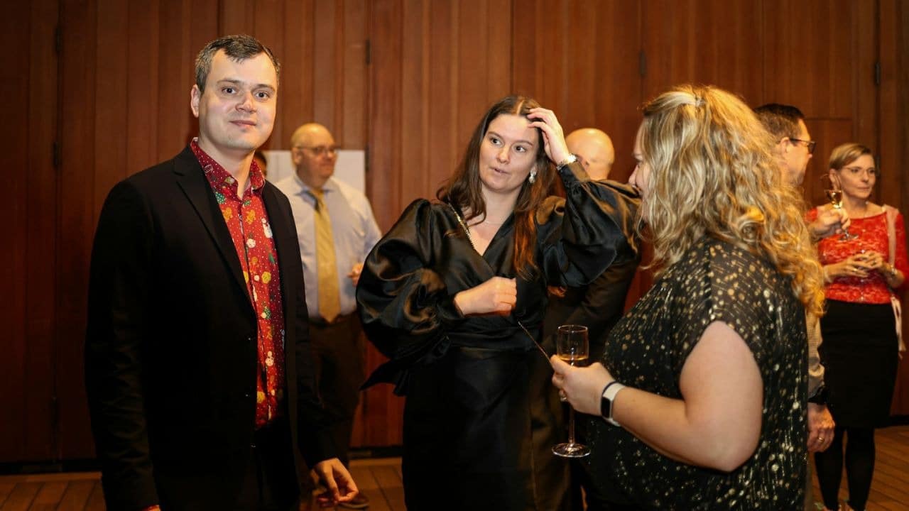 People in formal attire stand in a room, holding glasses and talking during a social event.