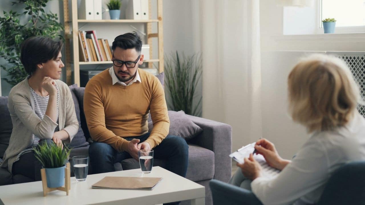 A man and woman sit on a couch facing a person holding a clipboard.
