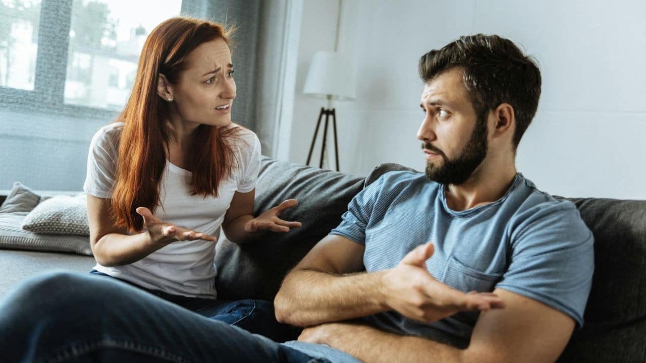 A woman gestures with open palms while talking to a man on a gray sofa.