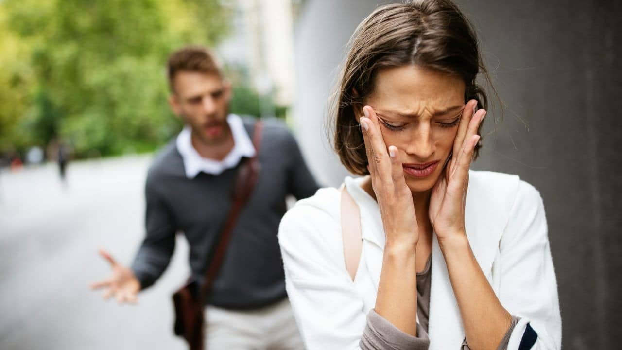 A woman covers her face with her hands while a man gestures in the blurry background.