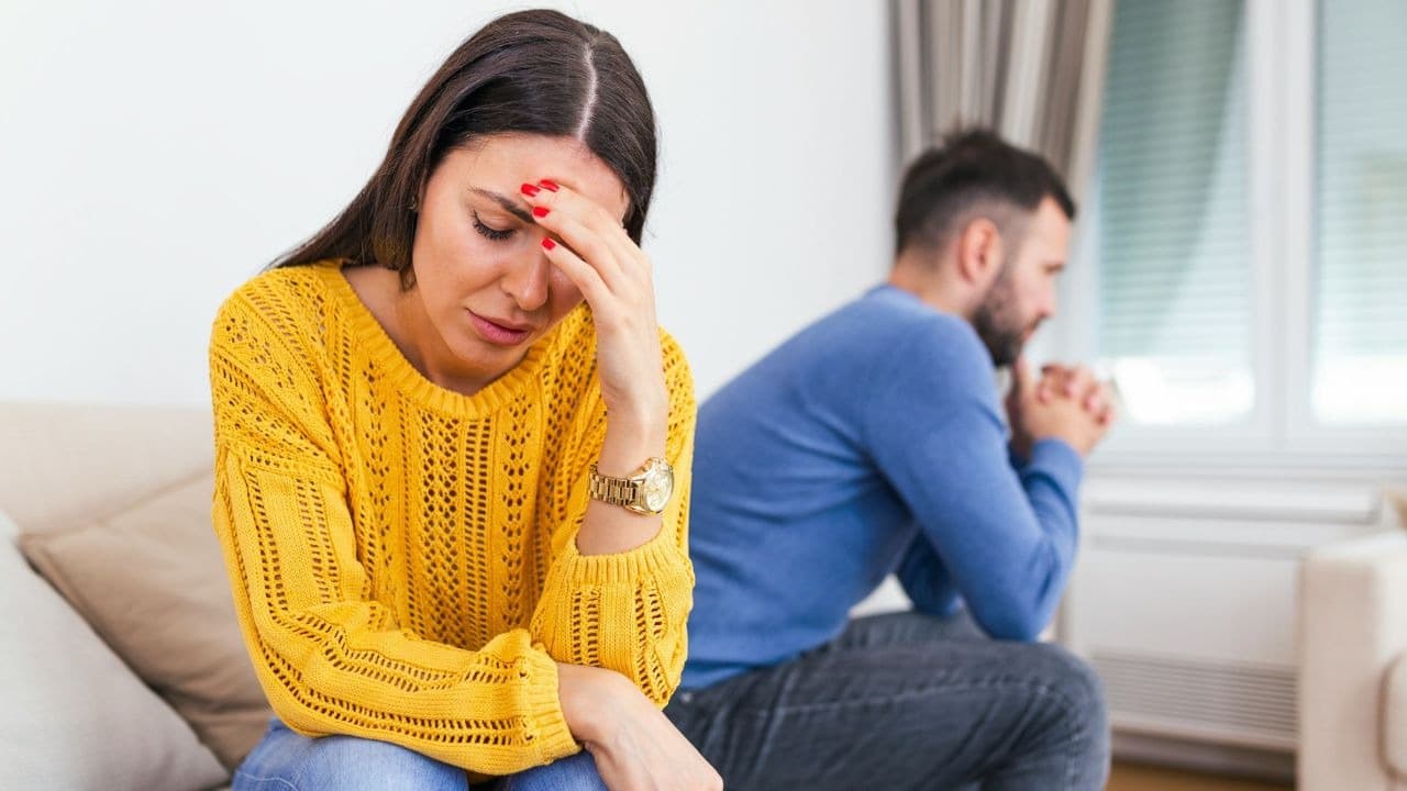 A woman in a yellow sweater touches her forehead while a man sits behind her.