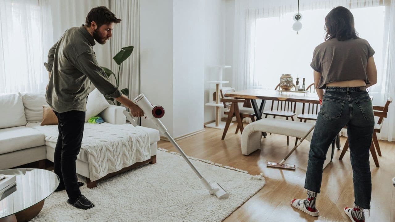 A man vacuums a rug while a woman mops the floor in a bright living room.