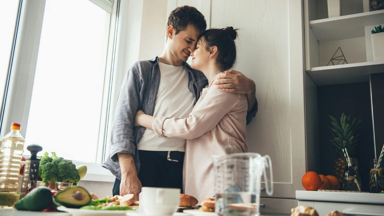 A man and woman embrace in a kitchen with their foreheads touching near a window.