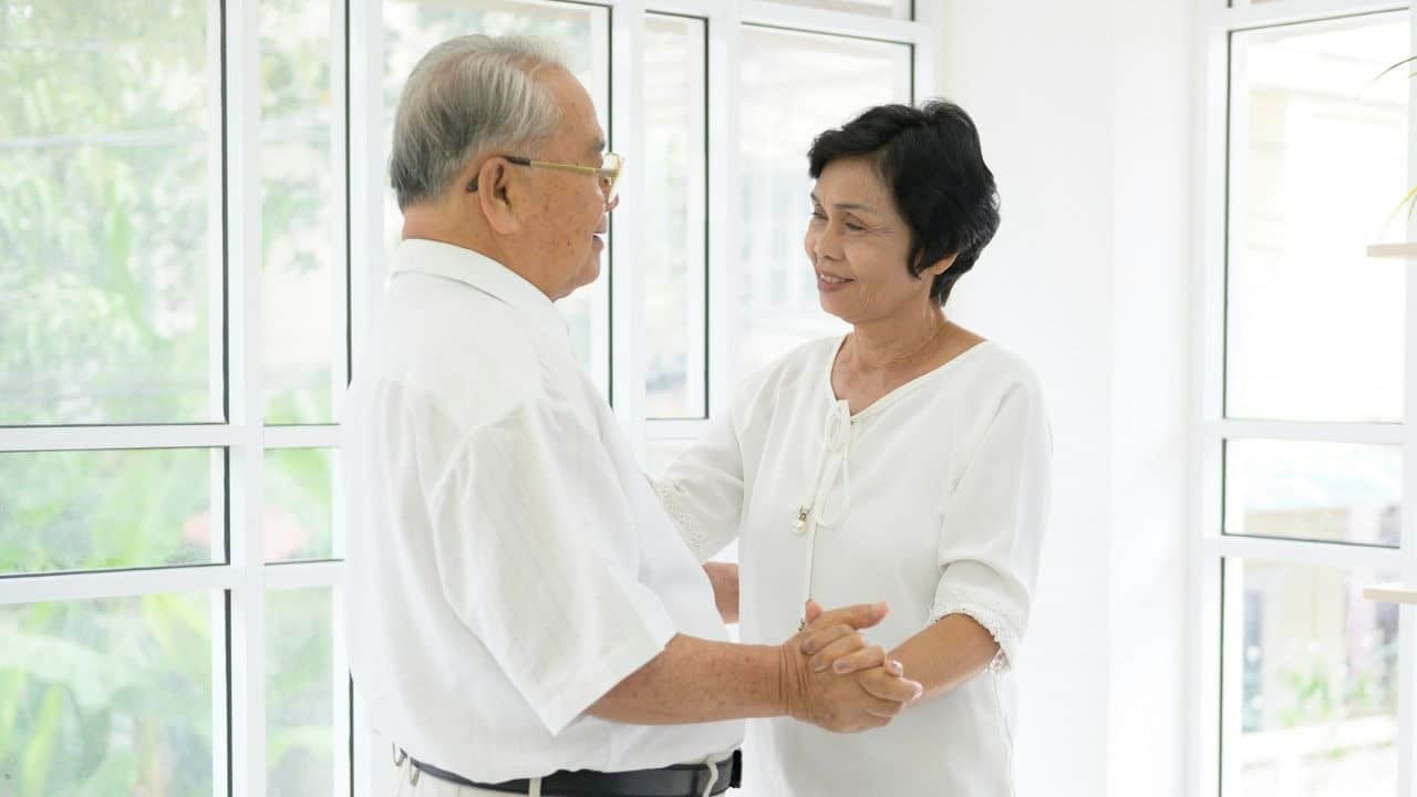 An elderly man and woman hold hands and smile while dancing in a bright white room.