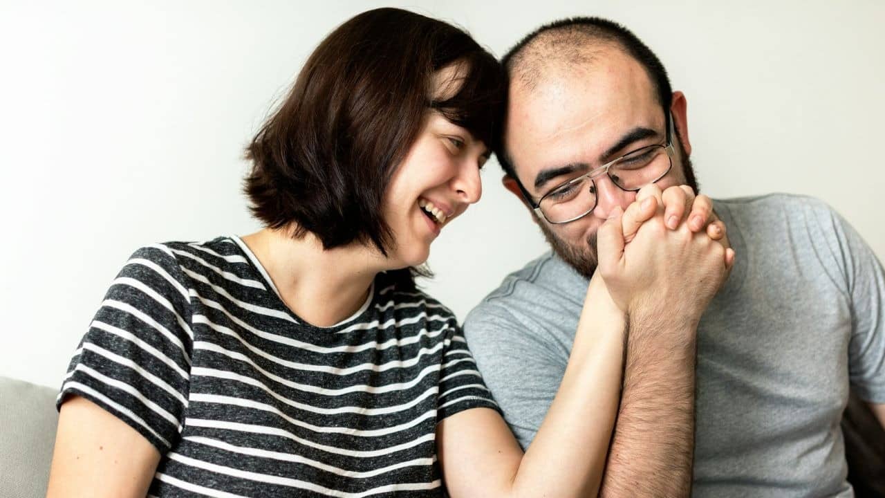 A man and woman laugh together while holding hands with their heads leaning toward each other.