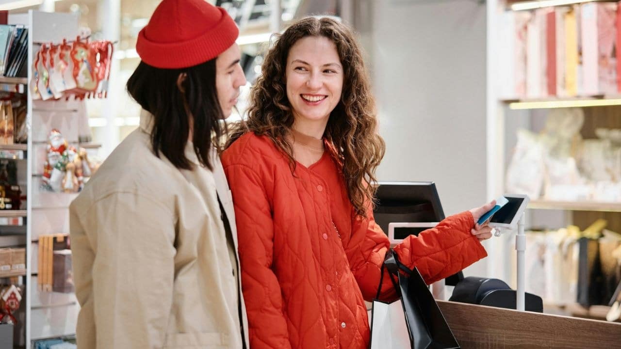 A smiling woman in a red jacket uses a card to pay at a store counter.