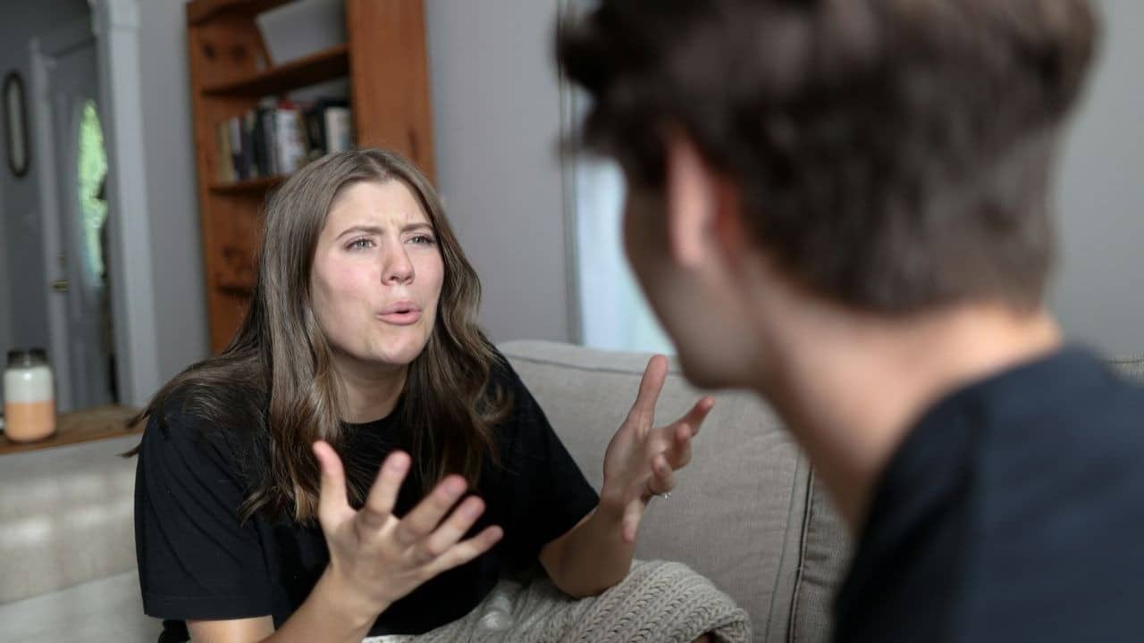 A woman with an upset expression gestures with her hands while speaking to a man.