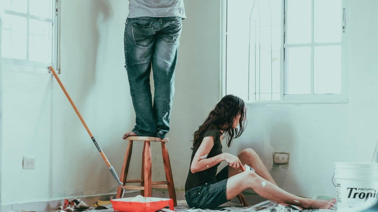 A woman paints a wall while a man stands on a stool nearby in a room.