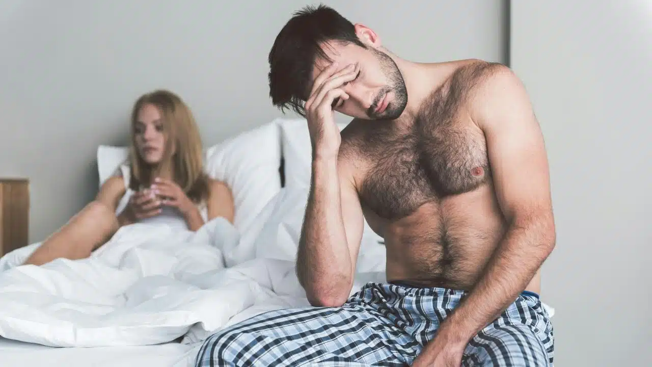 A shirtless man sits on a bed with his hand to his head, looking down.