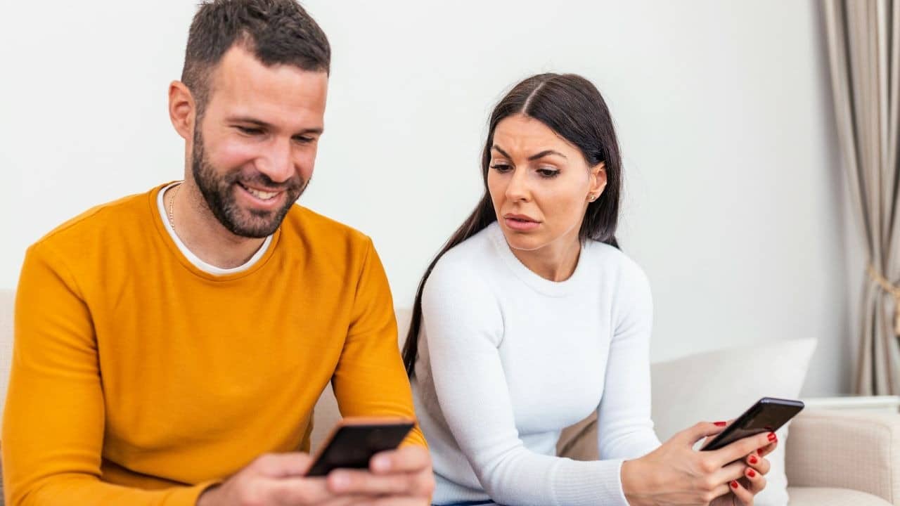 A woman looks suspiciously at a man's phone while they both hold their own devices.