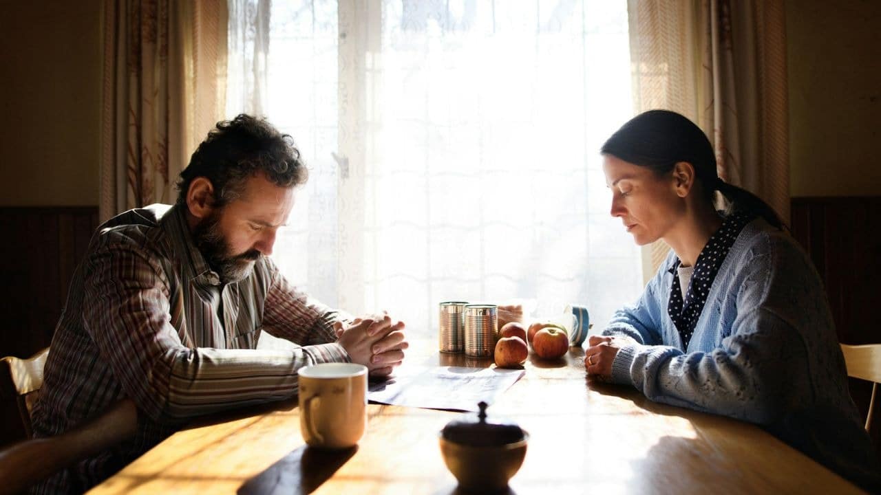A man and woman sit at a table with their heads bowed in a sunlit room.