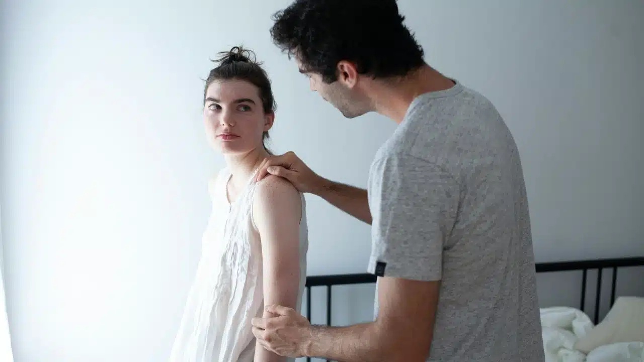 A woman sits on the edge of a bed while a man watches her from behind.