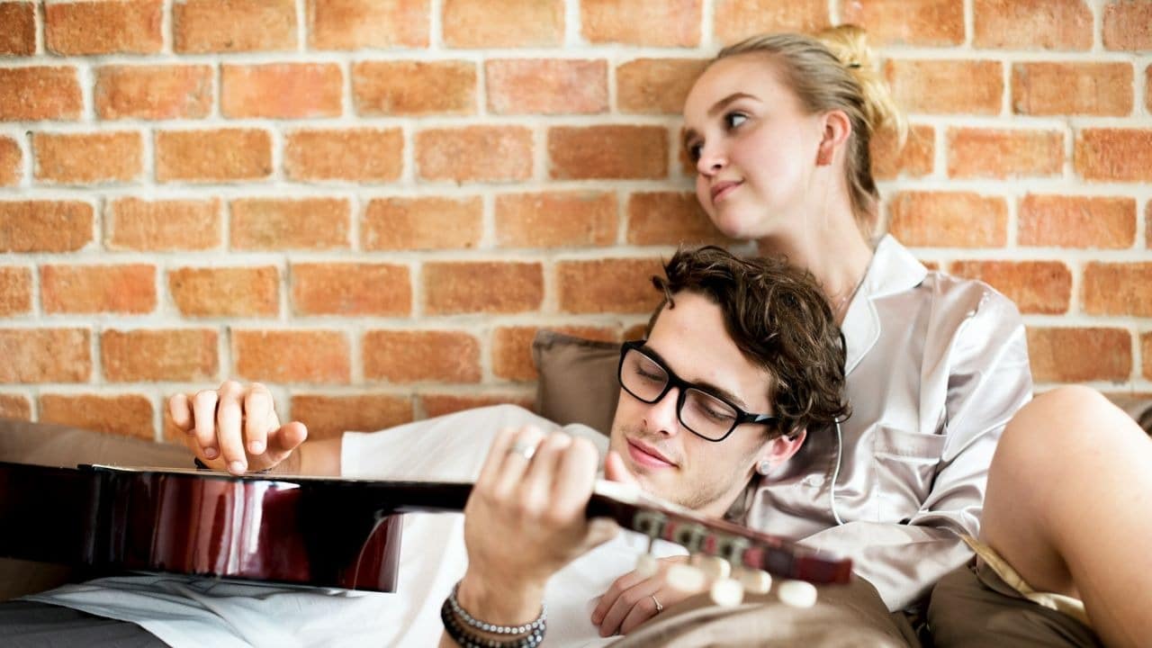 A man plays an acoustic guitar while a woman leans against him in front of brick.