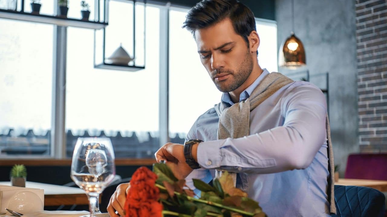 A man sits alone at a restaurant table with roses and looks at his watch.