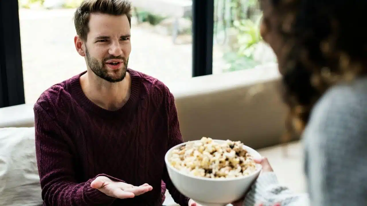A man in a purple sweater gestures toward a bowl of popcorn held by another person.