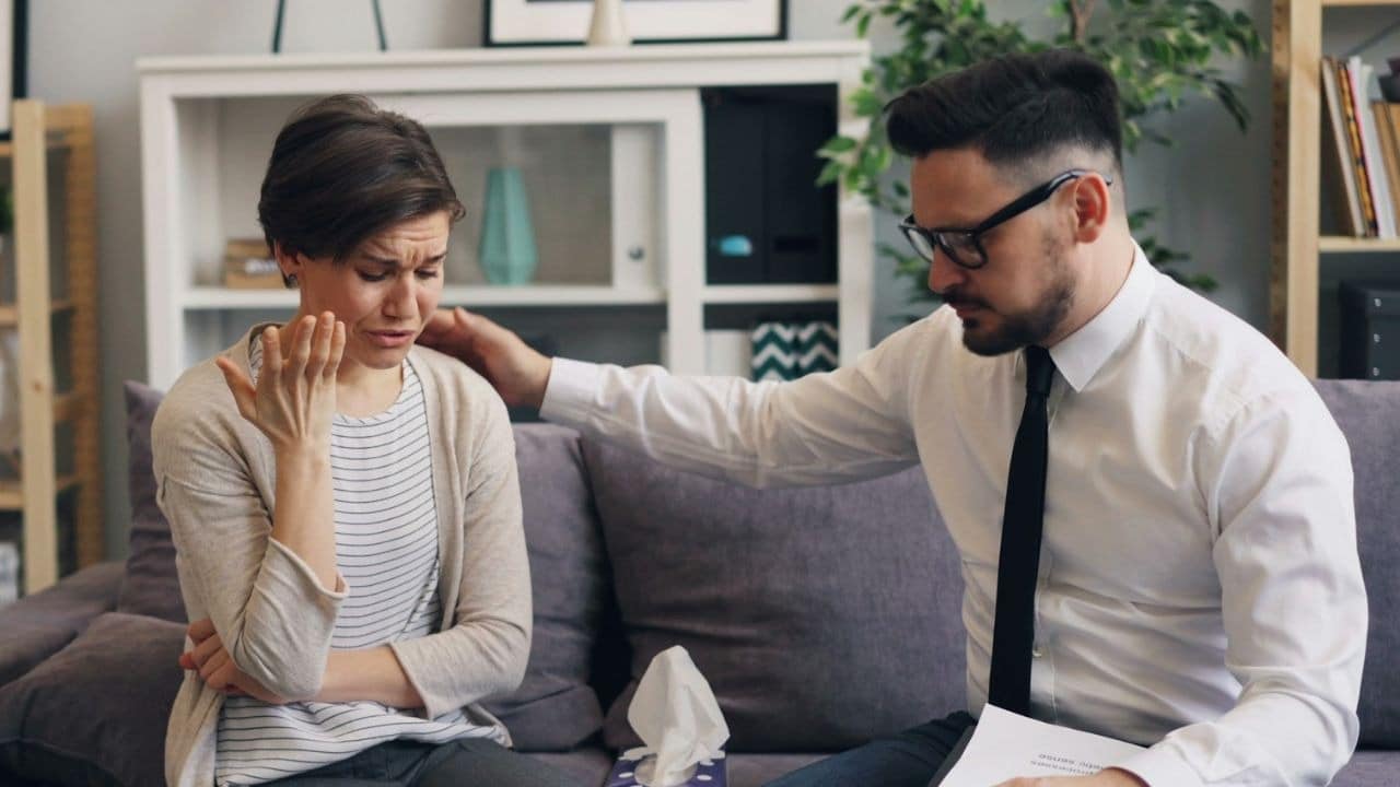 A man in a white shirt and tie comforts a crying woman on a couch.