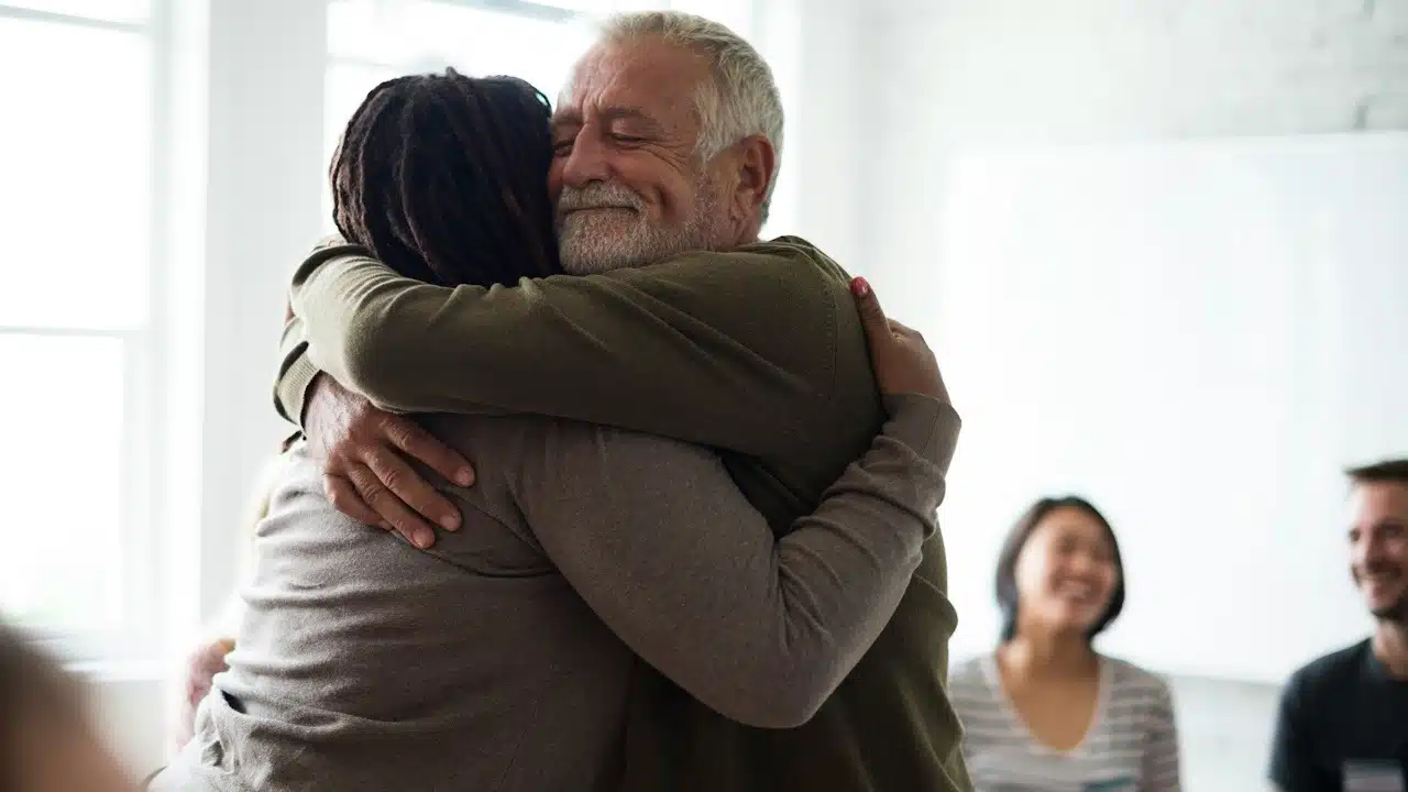 An older man hugging his wife while some people smile in the background.