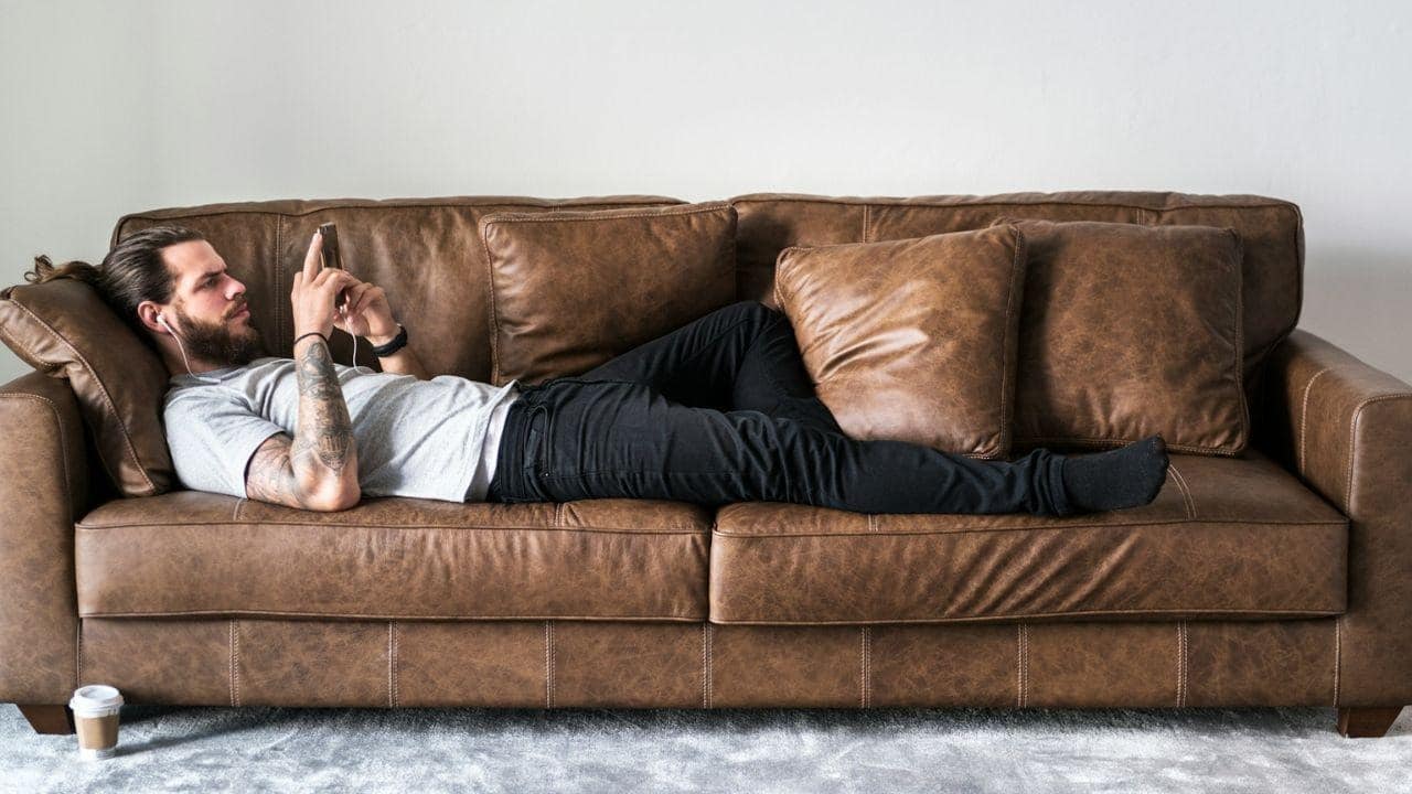 Bearded man with tattoos lying on a brown leather couch, looking at his phone.