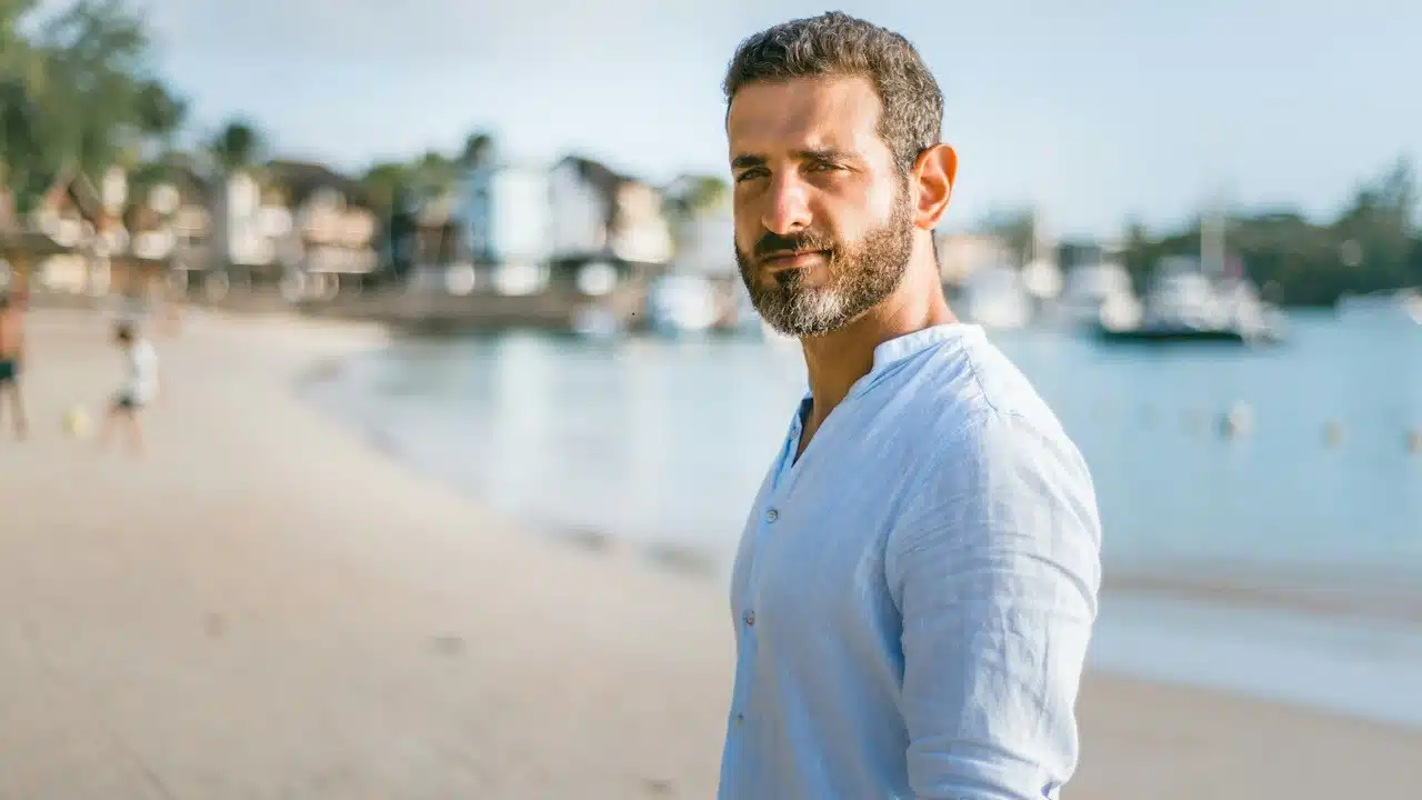 A bearded man standing at the beach and looking at the camera.