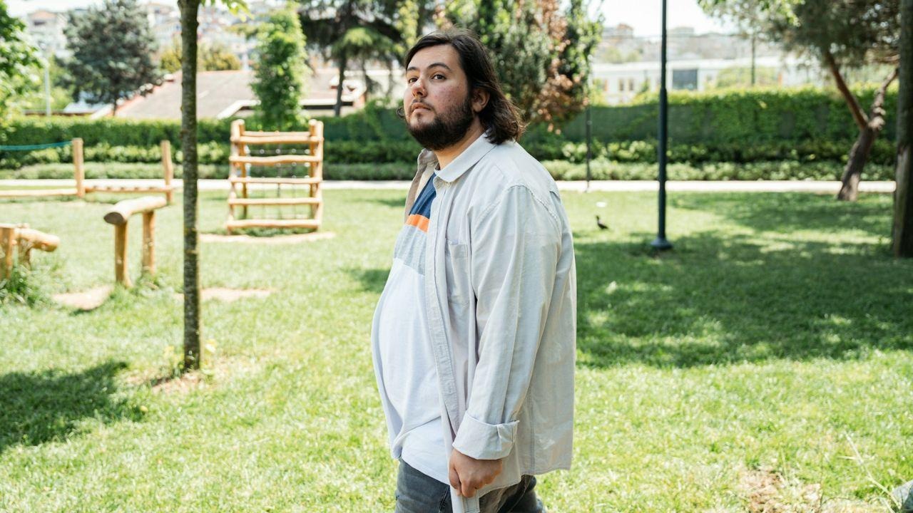 A man with a beard and long hair wearing an oversized shirt in a sunny park.