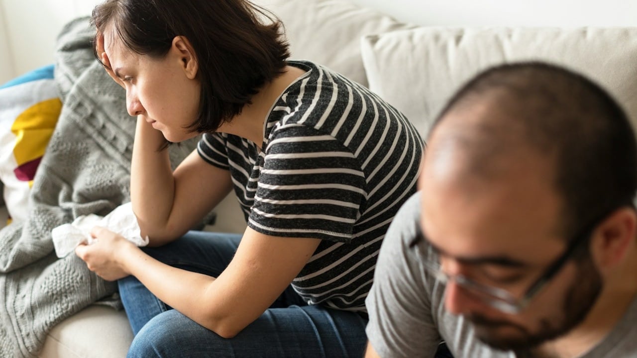 A woman holding her head in her hand while sitting on a couch while her husband looks away.