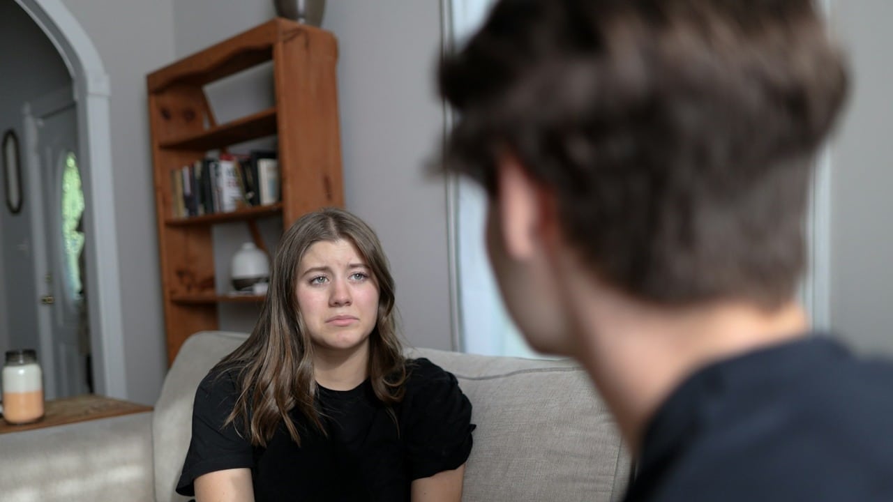 A woman looking at a man while sitting on a couch with a sad expression on her face.