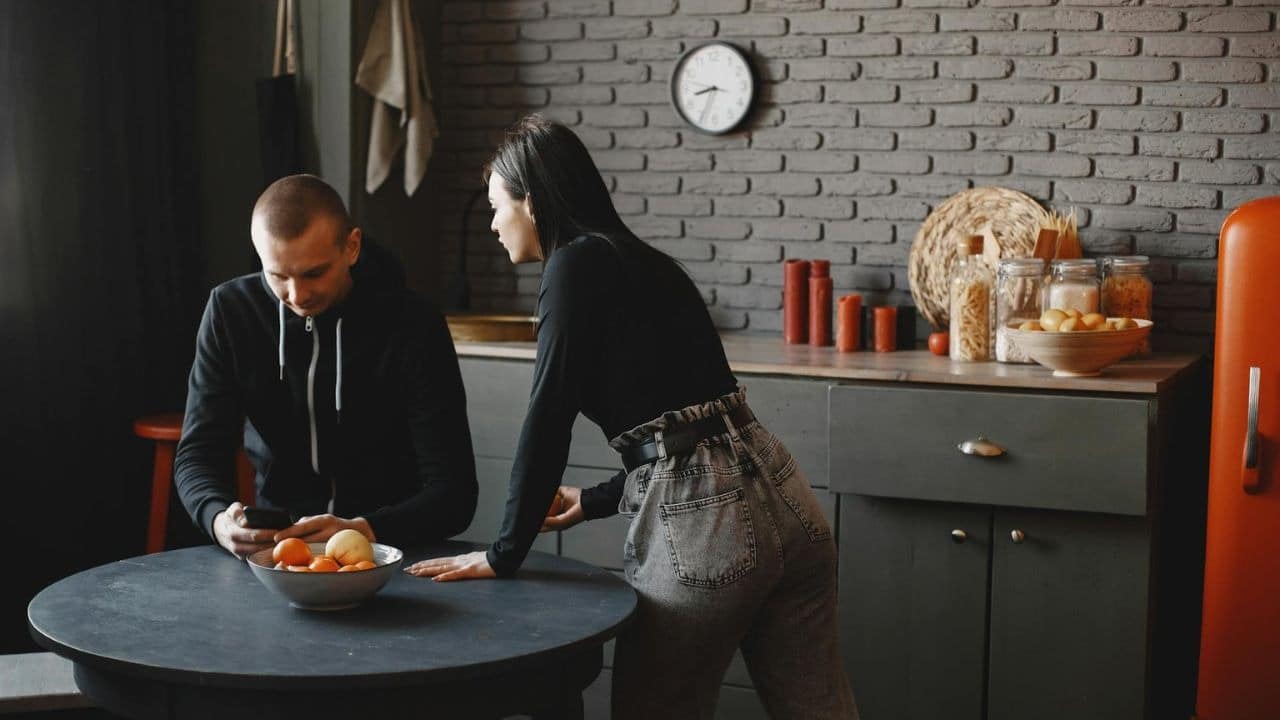 A woman stands in a kitchen looking at a man sitting and using a phone.