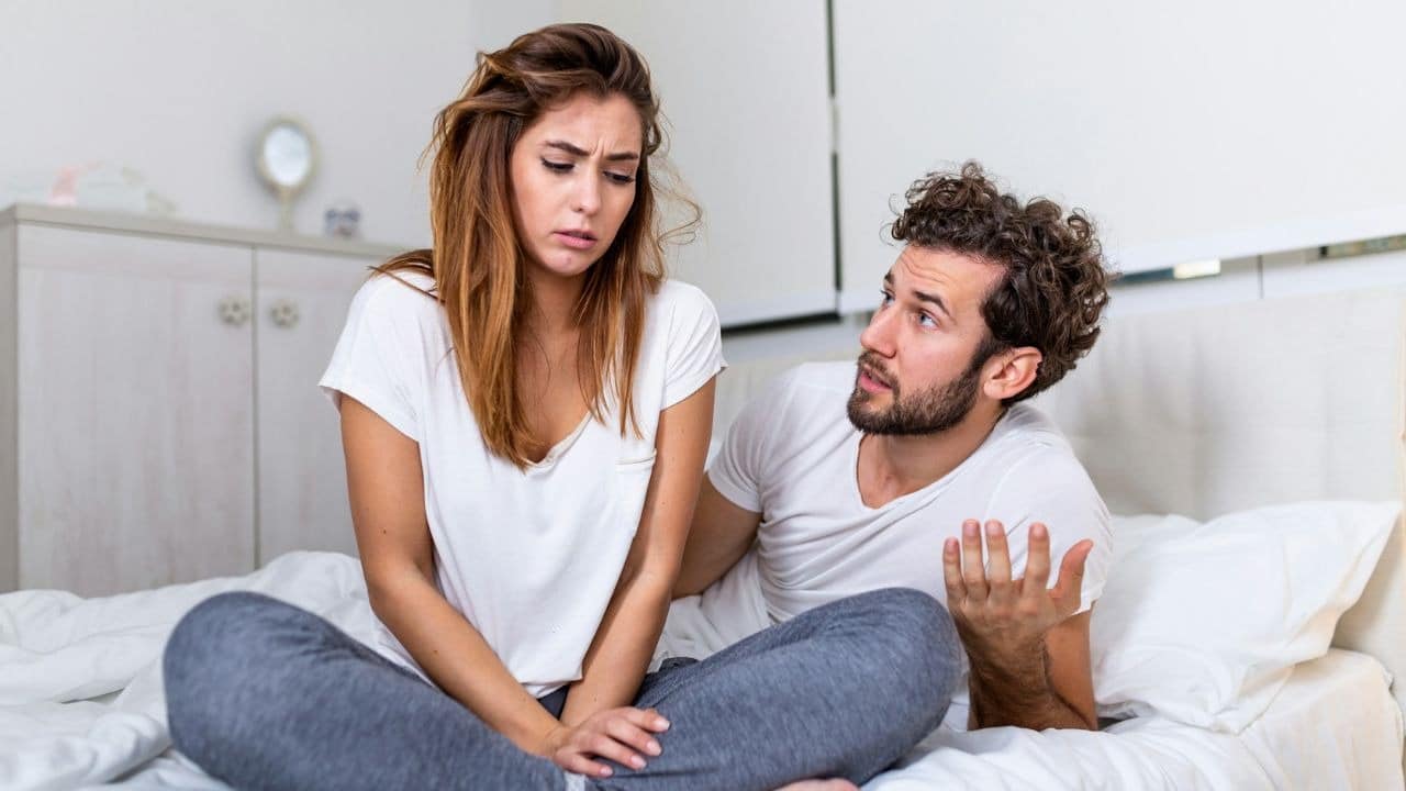 A worried woman sits on a bed while a frustrated man gestures and talks to her.