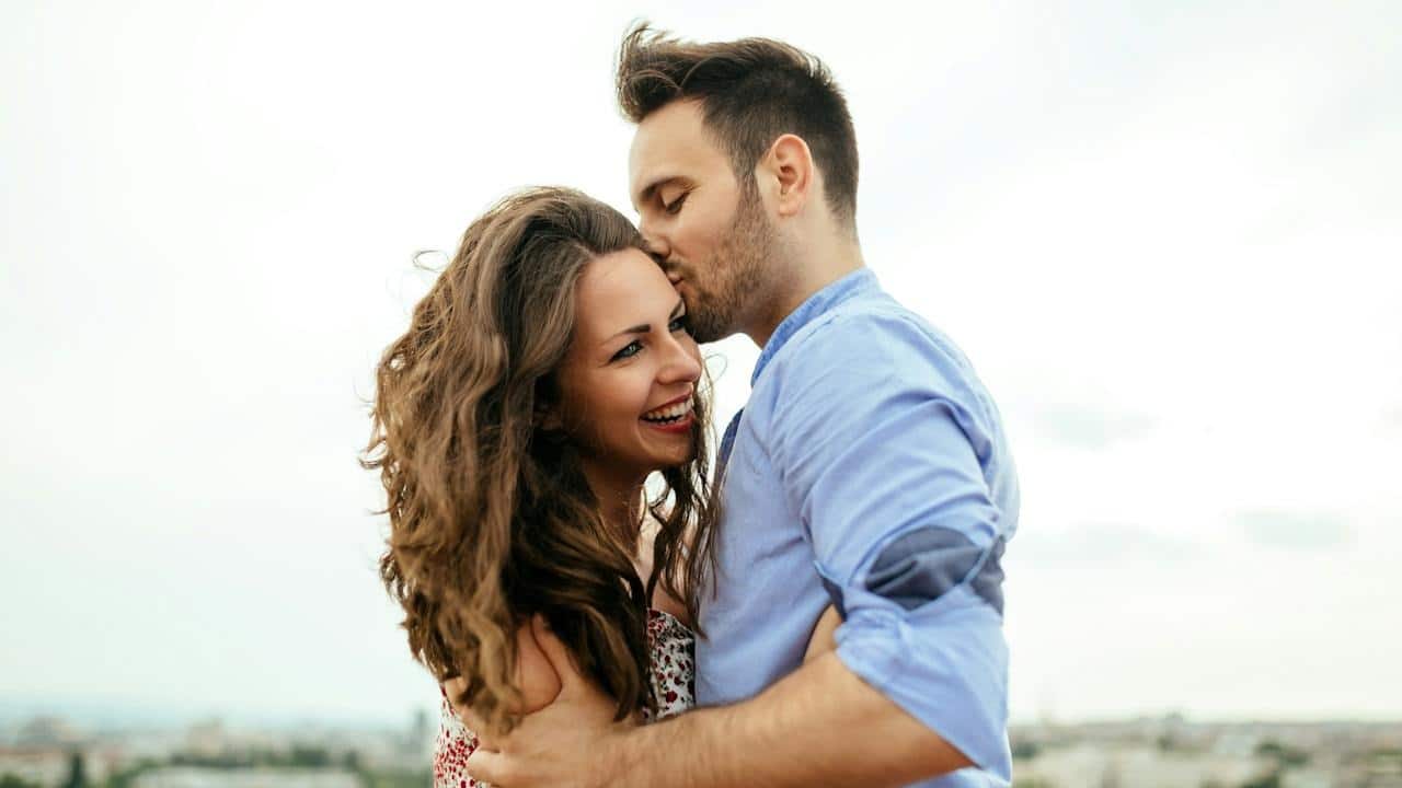 A lightly bearded man kissing a smiling woman on her temple.