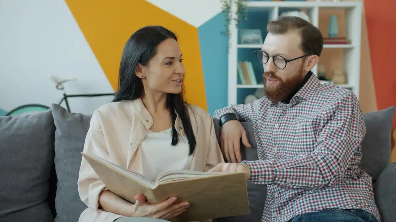 A woman showing a bearded man something in a book as they sit on a couch.