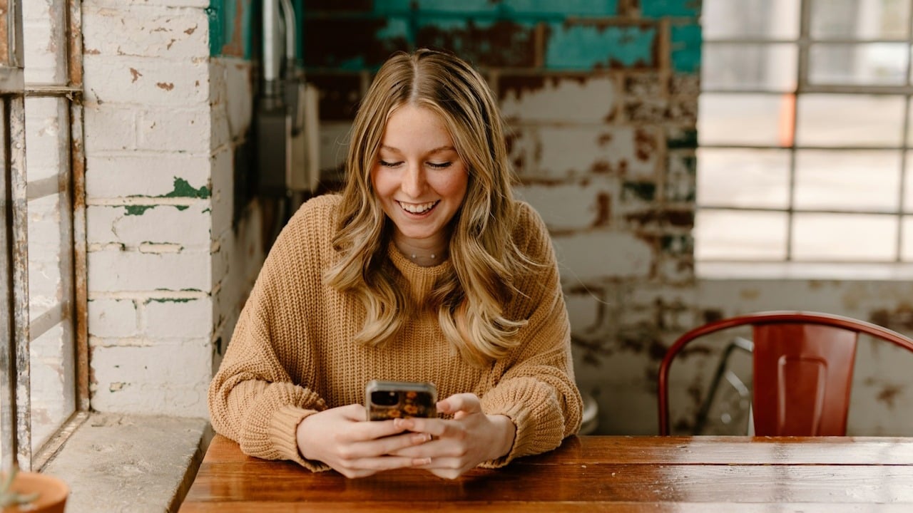 A blond woman smiling as she texts while sitting at a table.