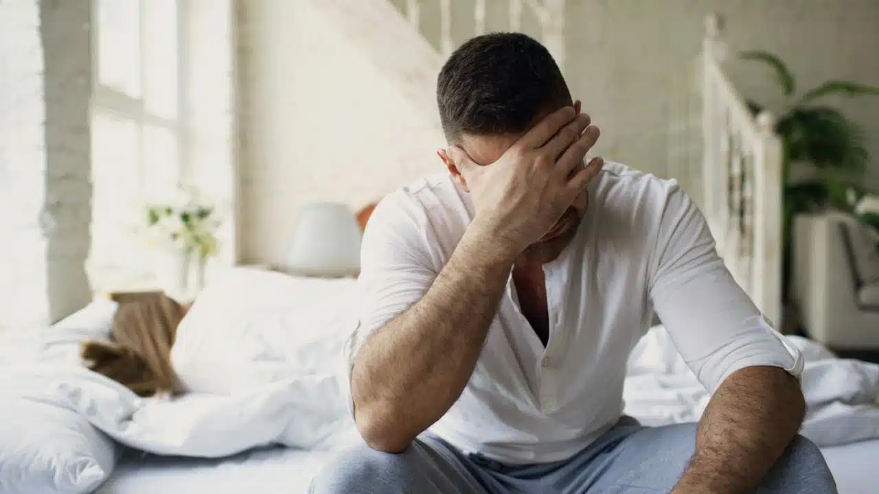A stressed man is sitting on a bed while another person is lying in the background under the covers.