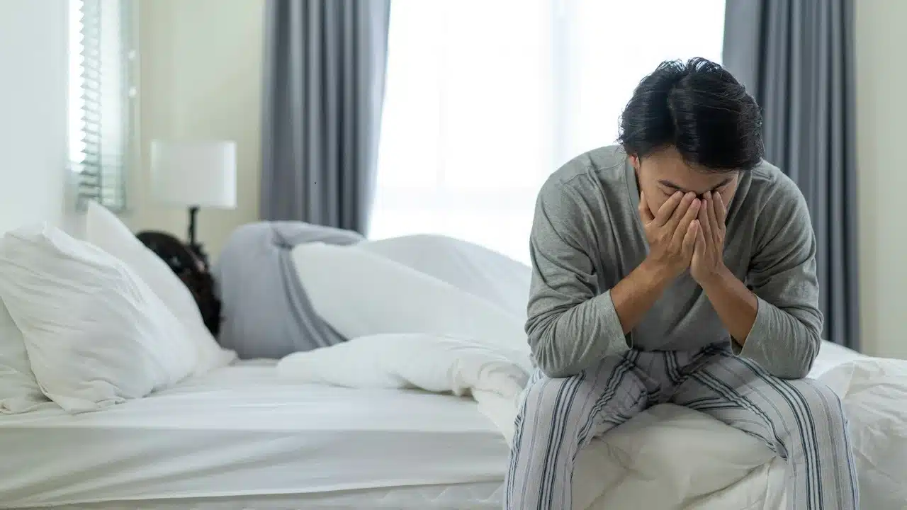 A man sitting on the edge of a bed with his face in his hands as his wife sleeps behind him on the bed.