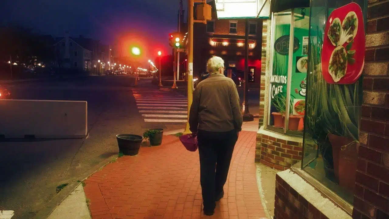 A person walks down a brick sidewalk at dusk, passing a cafe window and streetlights.