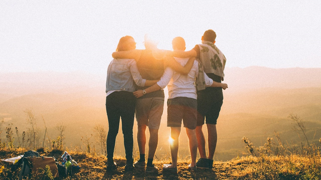 A group of friends standing together with arms on each other's shoulders as they stand outdoors in front of the sun on a cliff overlooking a scenic view.