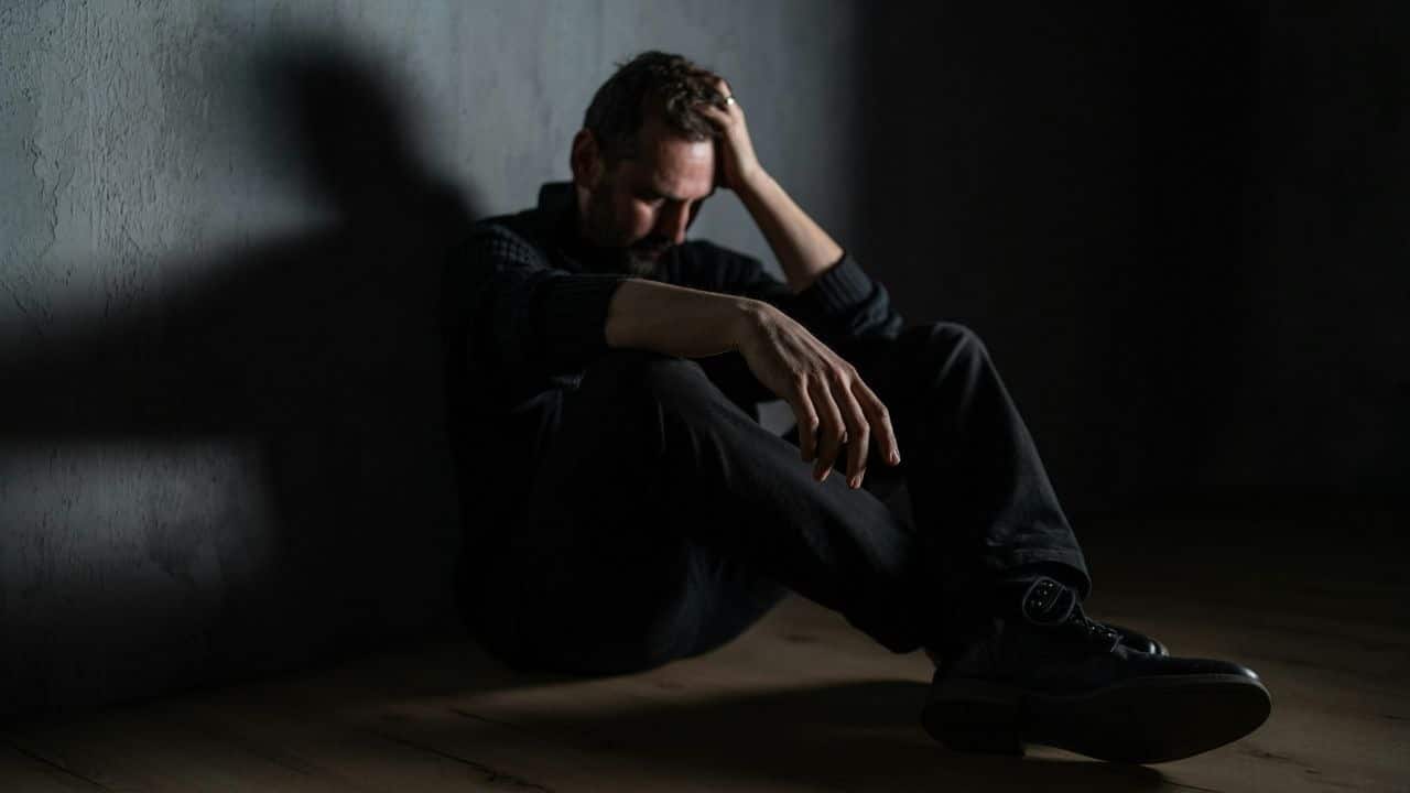 A man sits alone in a dark room with his hand on his head, looking stressed and sad.