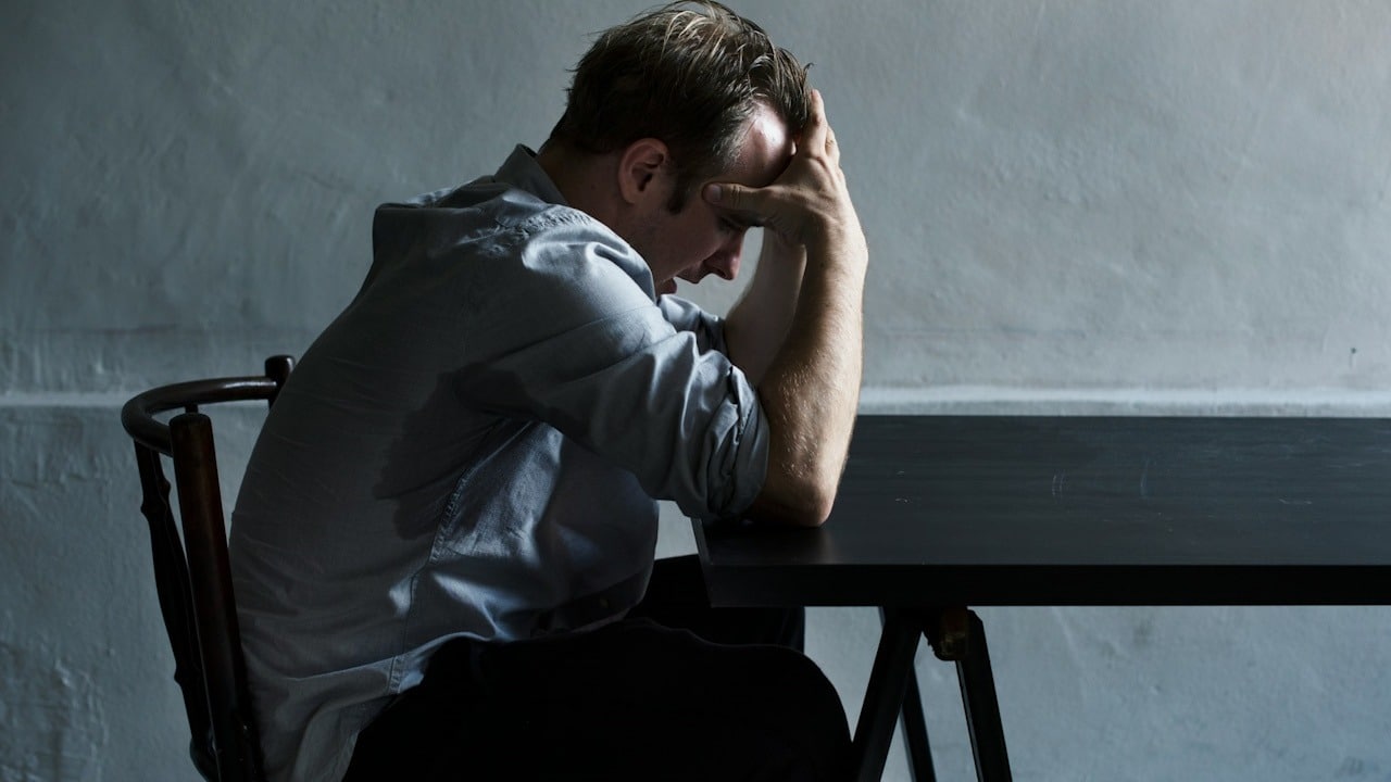 A worried man sitting at a table on a chair and holding his head in his hands.