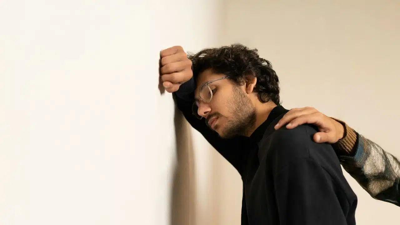 A man leans his head against a white wall while a hand rests on his shoulder.