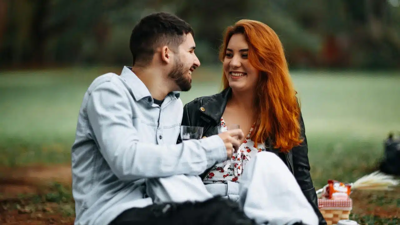 A man and a woman with red hair sit on the grass laughing during a picnic.©Matheus Bertelli/Pexels.com