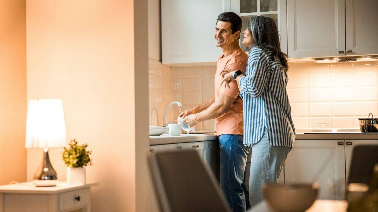 A couple smiles while washing dishes together in a bright, modern kitchen.