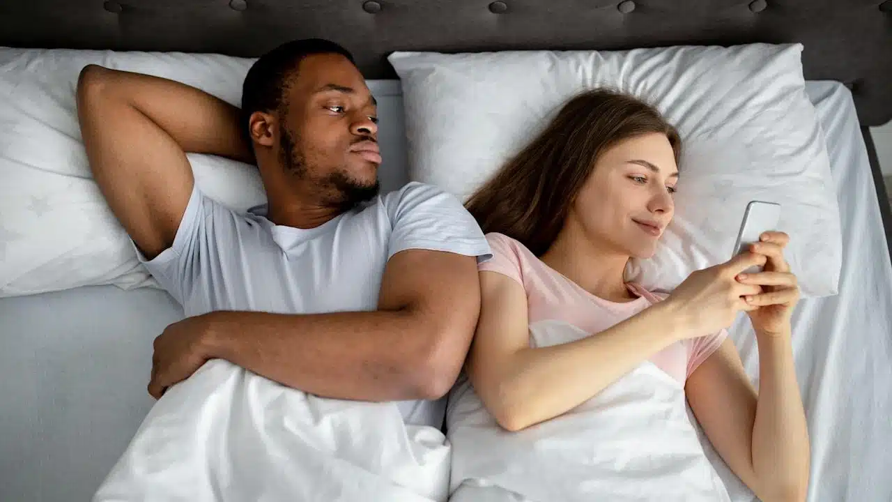 A man is lying in bed, looking suspiciously at the woman next to him who is smiling while looking at her phone.