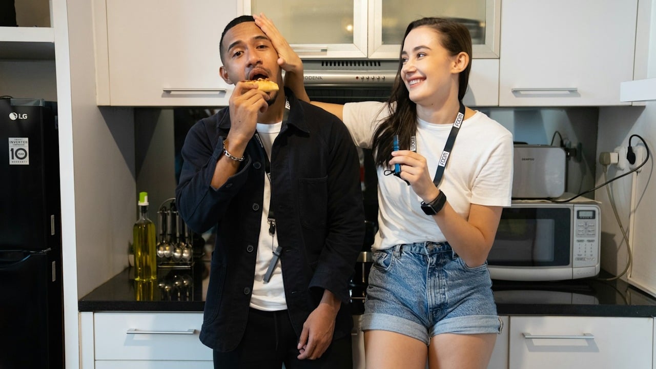 A woman playfully pushing a man’s head while he eats a slice of pizza as they lean against a cabinet in a kitchen.